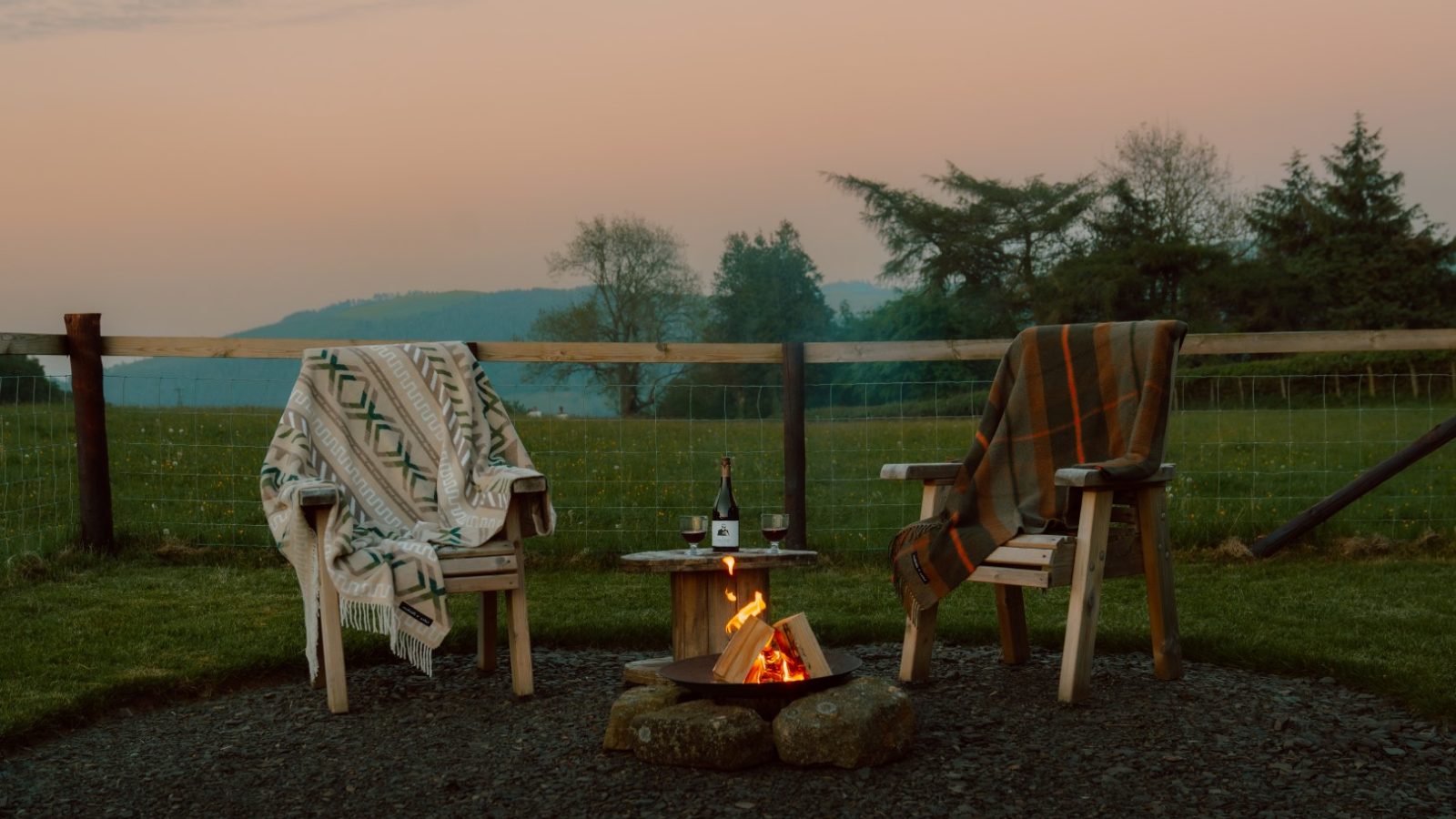 Two chairs with blankets by a small fire pit at Y Caban, enclosed by trees and a wooden fence, basking in the sunset glow.