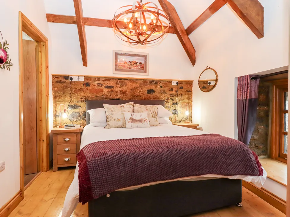 Cozy granary-themed bedroom with wooden beams, stone wall, double bed, red throw blanket, and warm pendant light.