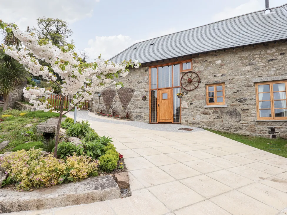 Bagtor Granary cottage with wooden door, wagon wheel, and blooming tree on a paved path surrounded by greenery and flowers.