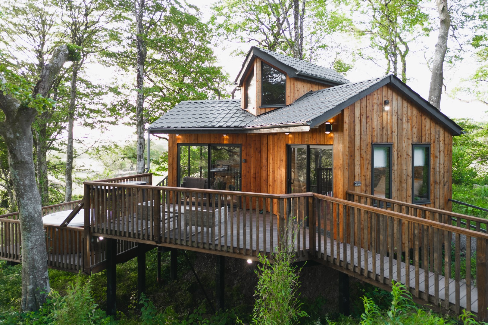 A wooden cabin with large windows and a deck is nestled among tall trees in the lush, green forest of the UK. #ToWander.