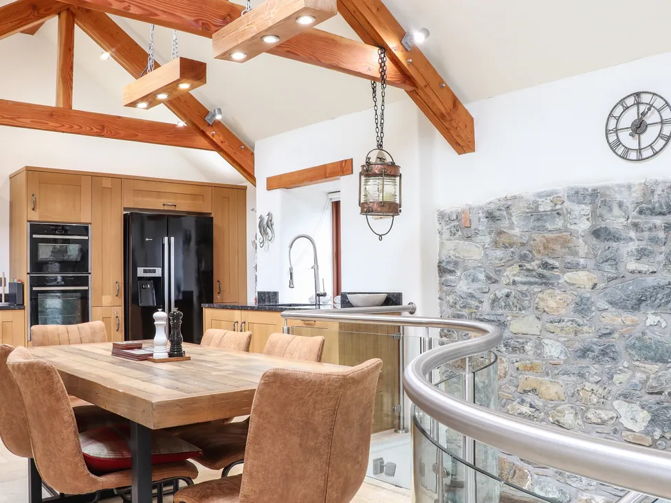 Modern kitchen at Pol Glas Hall with wooden beams, stone wall, dining table, and tan chairs under natural light.
