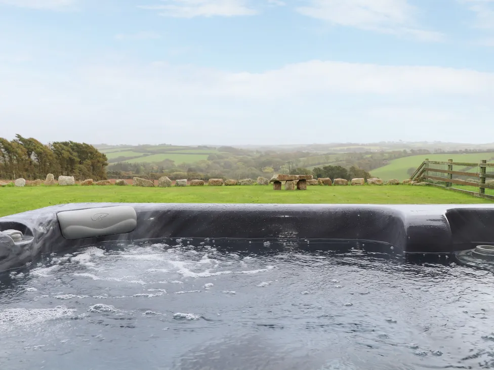 A bubbling hot tub at Pol Glas Hall overlooks a grassy field and rolling hills under a partly cloudy sky.