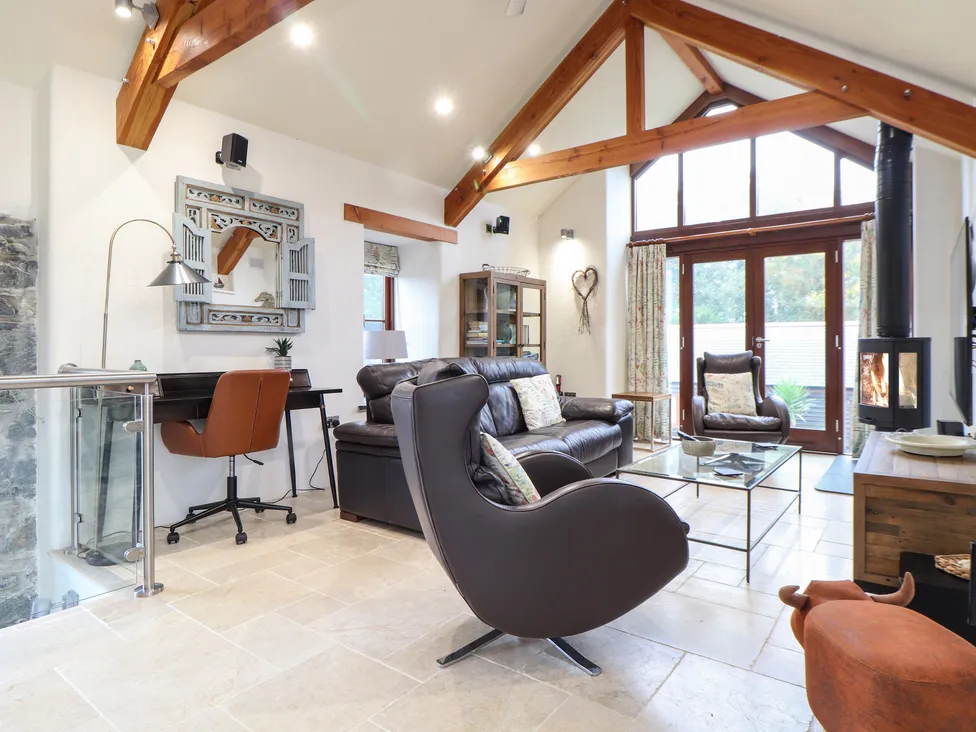 Modern living room at Pol Glas Hall with exposed beams, leather sofas, armchairs, and a wood-burning stove by large windows.
