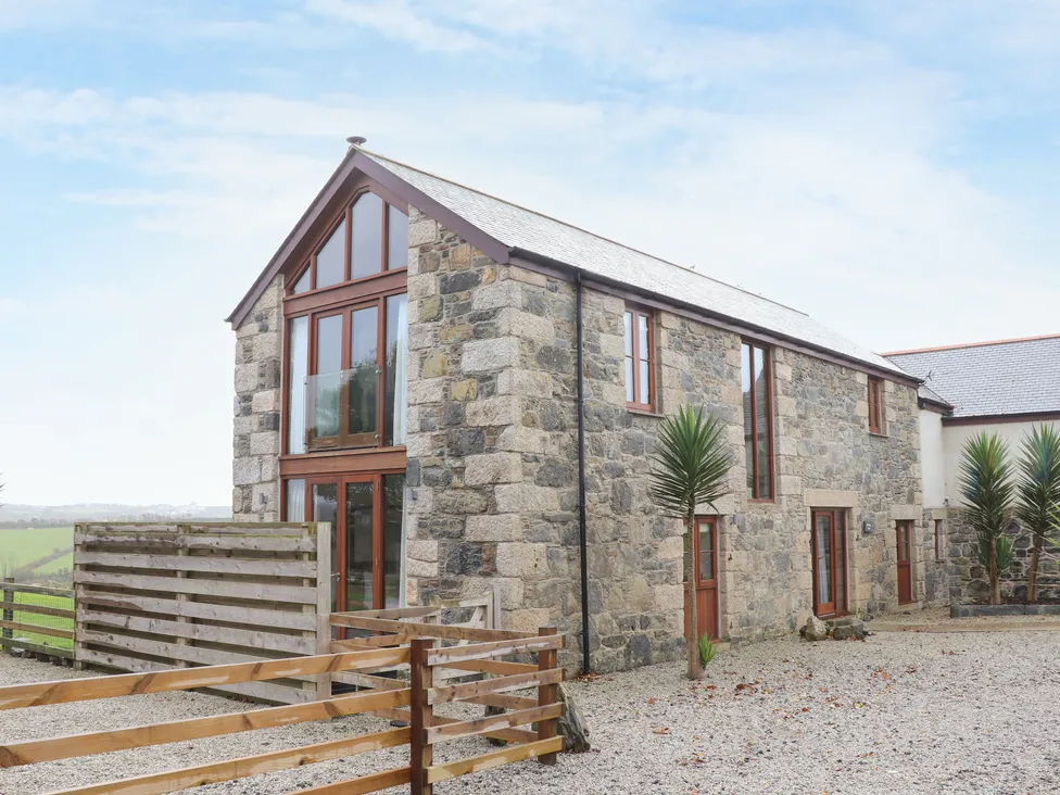 Stone barn, large glass windows, wooden fence, and palm trees under a blue sky at Pol Glas Hall.