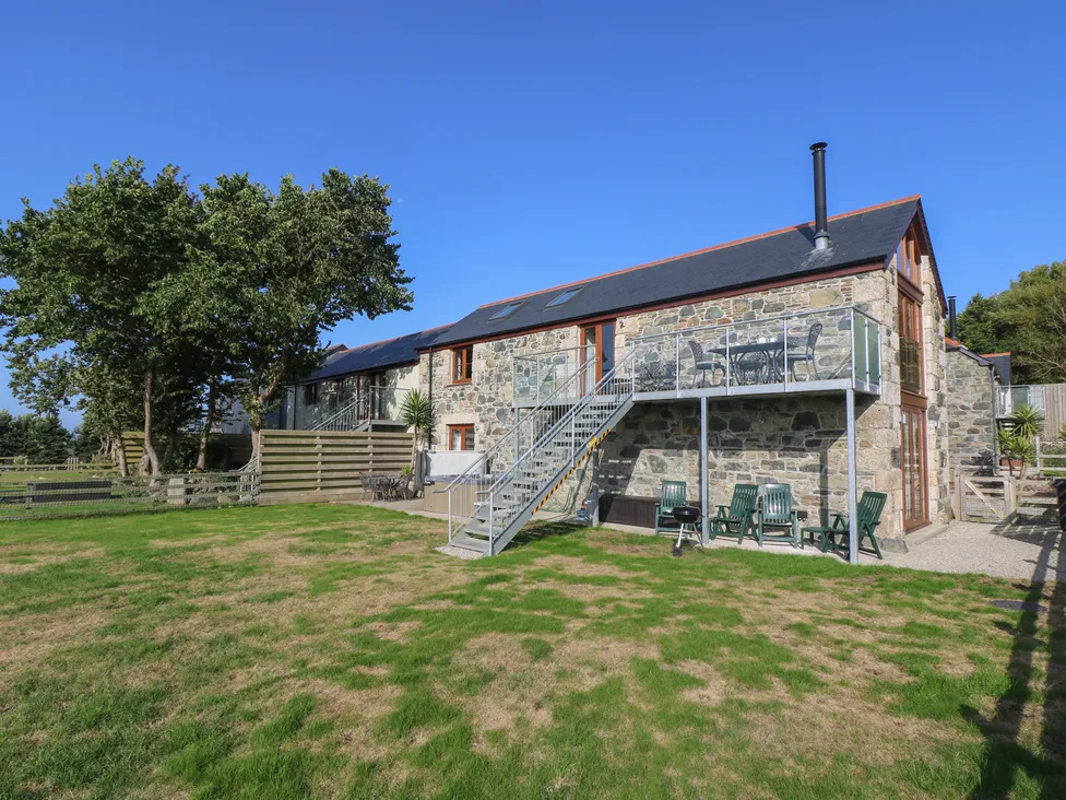Stone house at Pol Glas Hall with outdoor stairs, balcony seating, green lawn, and trees under a clear blue sky.
