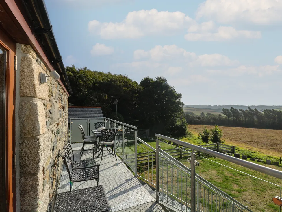 Metal balcony with chairs and tables at Pol Glas Hall overlooks fields, trees, and distant hills under a blue sky.