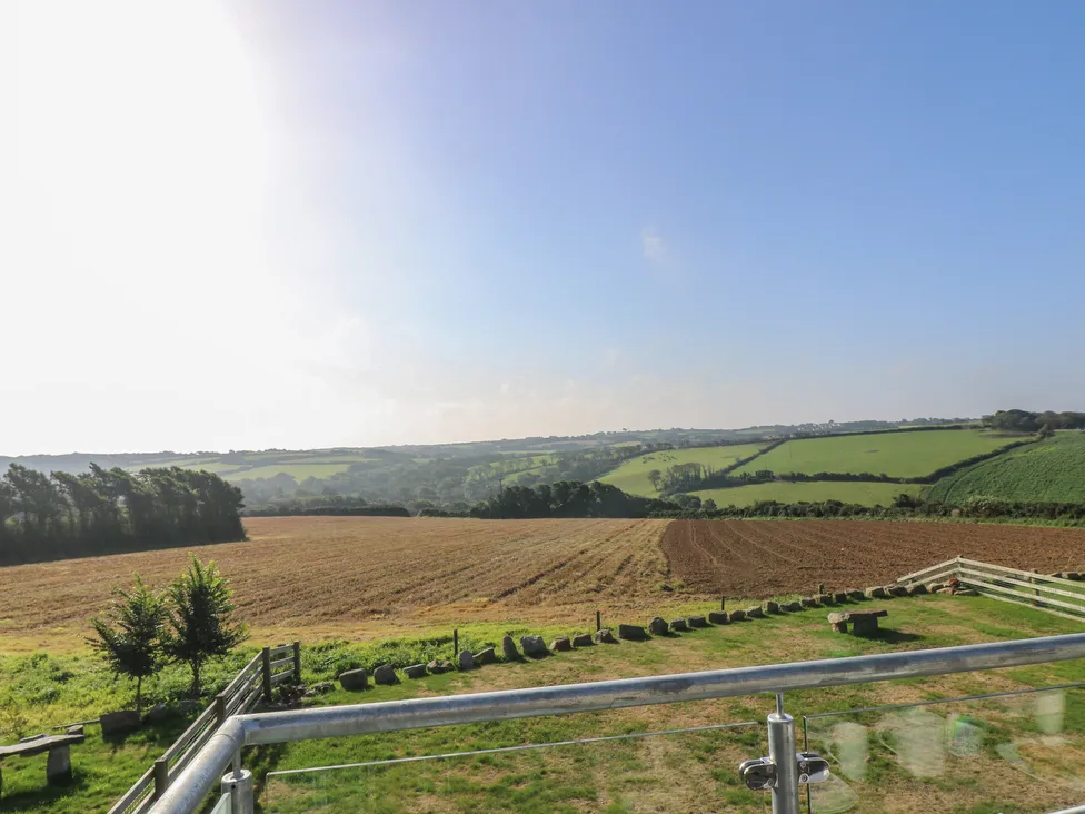 View of a sunlit countryside field near Pol Glas Hall, with green hills, trees, and a clear blue sky from a fenced terrace.