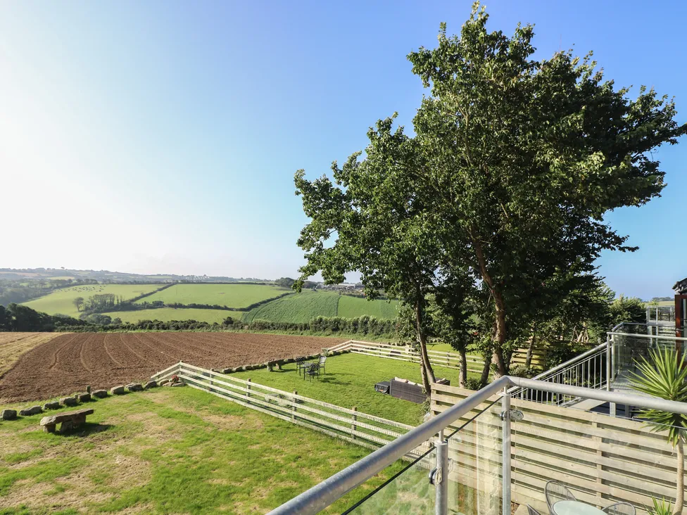 View from Pol Glas Hall balcony overlooking green fields, a plowed field, trees, and fenced grassy areas under a blue sky.