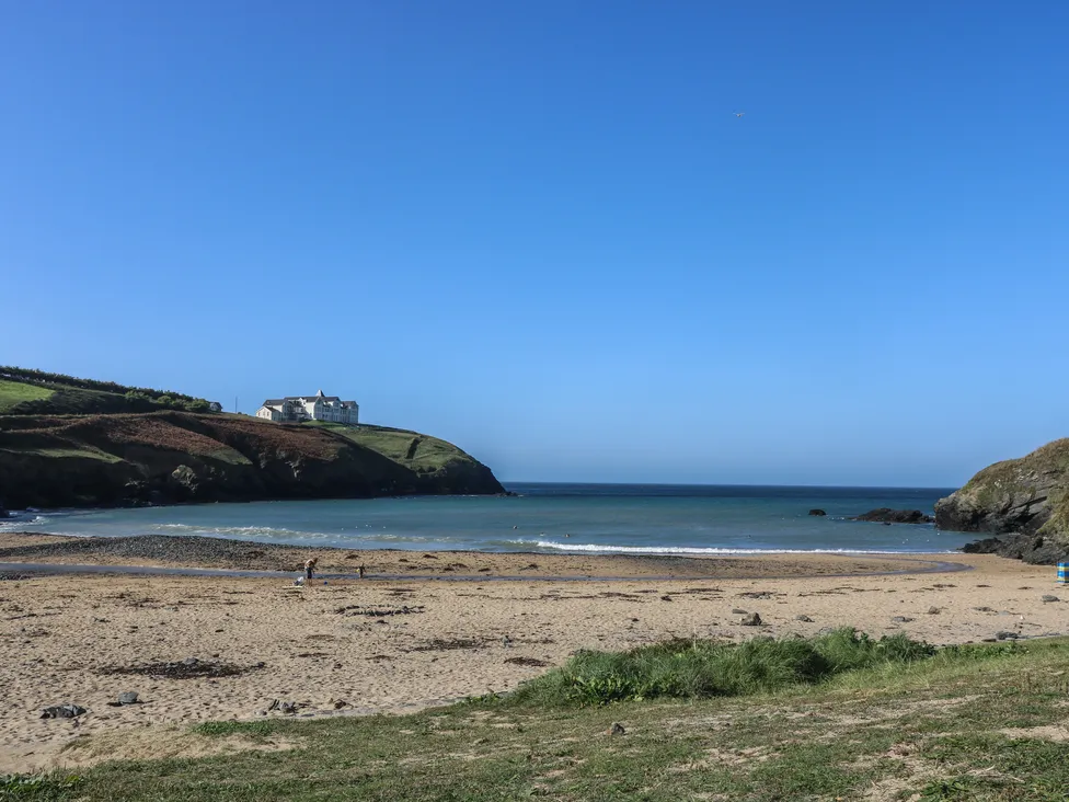 Sandy beach with gentle waves, grassy cliffs, and Pol Glas Hall visible under a clear blue sky.
