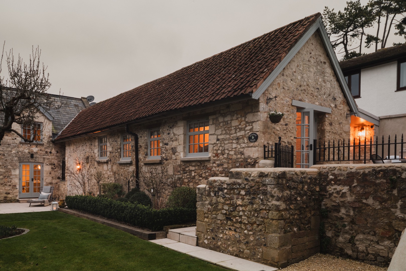 Stone cottage with warm, lit windows, surrounded by a tidy garden and lawn, under an overcast sky.
