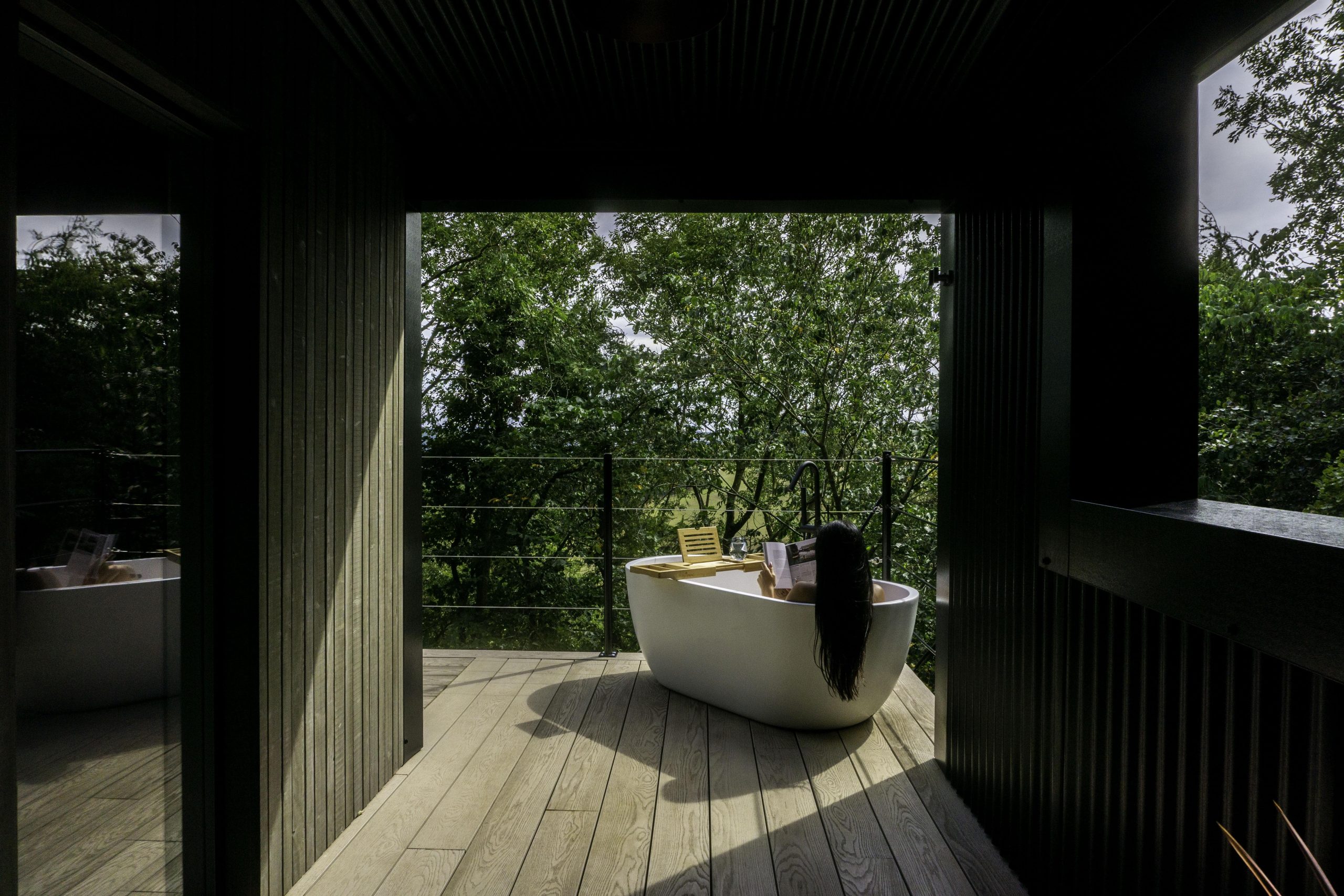 Woman reads a book in a freestanding bath with trees in the background at Bracken Treehouse