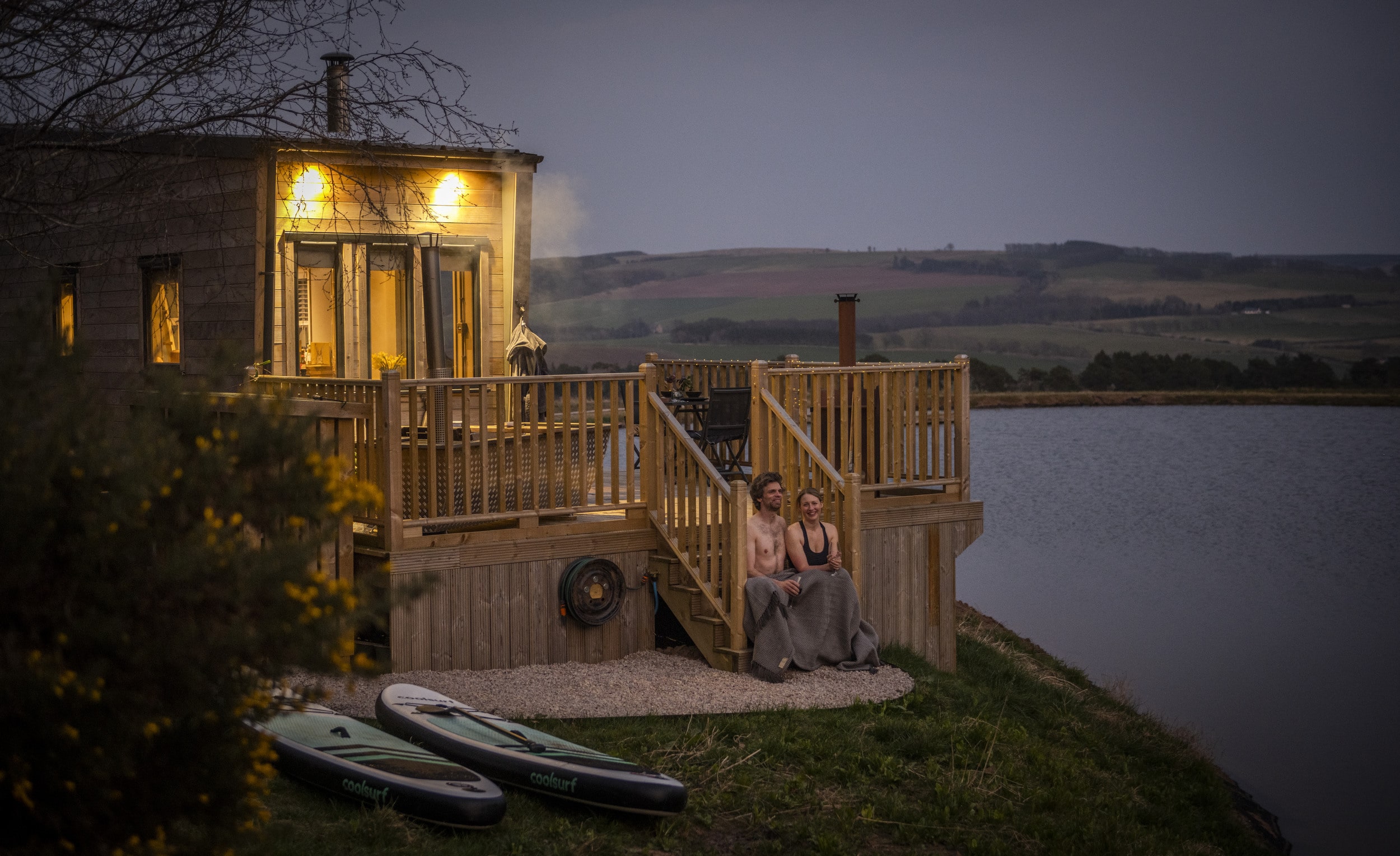 A couple cosy up in a blanket next to the lake at Tiny Home Borders cabin in Scotland